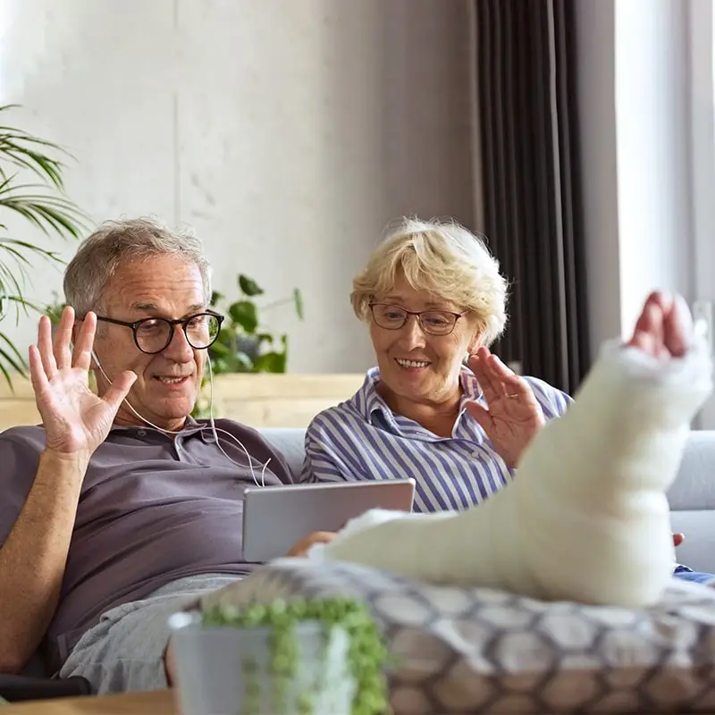 A cheerful senior couple sitting on a sofa and having a video call. The senior man has a broken leg.